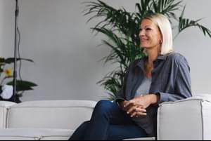 Maria Höfl-Riesch sitzt seitlich auf der Couch in einem Studio, verschränkt die Finger und lächelt.