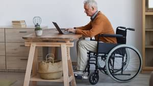 Ein älterer Mann im Rollstuhl sitzt am Schreibtisch auf dem sein Laptop steht