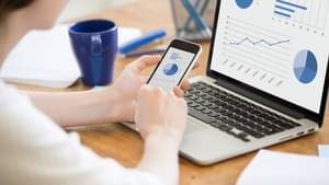 Eine Frau schaut auf ihre Finanzen auf dem Handy und einem Laptop auf dem Schreibtisch