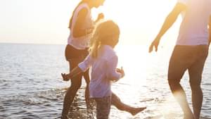 Eine Frau, ein Mann und ein kleines Kind laufen im Sonnenuntergang durch knöchelhohes Wasser und haben Spaß.